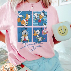 May include: Pink t-shirt with a cartoon image of Donald Duck in four different poses. The shirt has the text "Donald Duck" written in a cursive font below the image.
