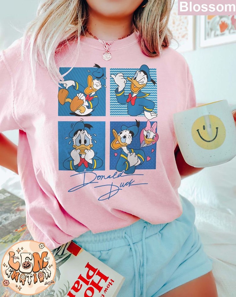 May include: Pink t-shirt with a cartoon image of Donald Duck in four different poses. The shirt has the text "Donald Duck" written in a cursive font below the image.