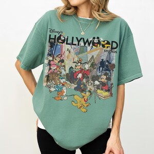 May include: A green t-shirt with a graphic print of Disney characters, such as Mickey Mouse, Minnie Mouse, and Donald Duck, with the text "Disney's Hollywood Studios". The shirt has a loose fit and short sleeves.