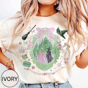 May include: Ivory-colored sweatshirt featuring a whimsical design with a green and pink framed image of a castle and two figures. The design includes a witch's hat, a crown, a broom, and the text "Wicked" and "Changed for Good."