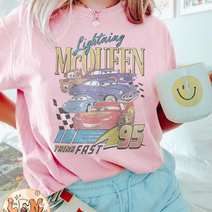 May include: Pink t-shirt with a graphic of Lightning McQueen from the Cars movie. The graphic includes the text "Lightning McQueen" and "Think Fast 95".