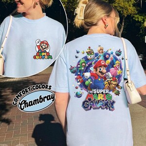 Pu&ograve; includere: T-shirt azzurra con grafica Super Mario Galaxy sul retro e grafica Super Custom Mario sul davanti. La maglietta &egrave; etichettata "Comfort Colors Chambray". La grafica sul retro presenta Mario, la Principessa Peach e altri personaggi.
