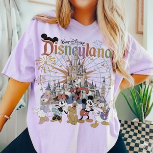 May include: A light purple t-shirt featuring a Disneyland graphic with Mickey Mouse, Minnie Mouse, Donald Duck, and other Disney characters. The graphic includes a castle, fireworks, and a Ferris wheel.