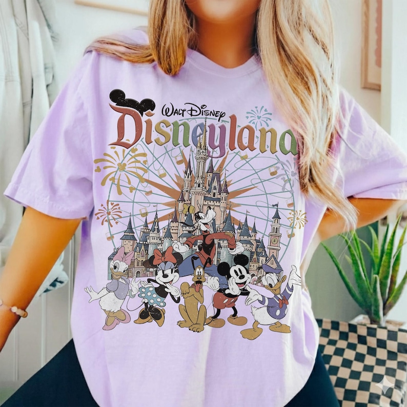 May include: A light purple t-shirt featuring a Disneyland graphic with Mickey Mouse, Minnie Mouse, Donald Duck, and other Disney characters. The graphic includes a castle, fireworks, and a Ferris wheel.