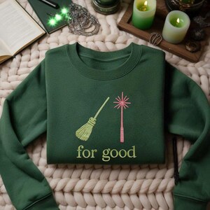 May include: Dark green sweatshirt with embroidered design of a broom and a wand, with the words "for good" below. The broom is light green, and the wand is pink. The sweatshirt is folded on a cream-colored, textured surface.