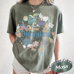 May include: Olive green t-shirt with a Disneyland graphic. The design features various park icons, a rainbow, and the word "Disneyland" in blue. The shirt also has the words "Comfort Colors" and "Moss" printed on it.