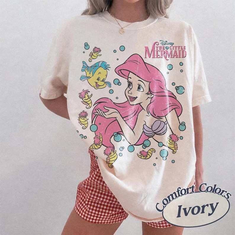 May include: An ivory short-sleeve t-shirt featuring a graphic print of Ariel from The Little Mermaid with pink hair, a purple seashell top, and Flounder. The design includes blue bubbles and the text Disney The Little Mermaid in pink lettering.