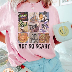 May include: Pink t-shirt with a graphic of a collage of various Disney characters celebrating Halloween. The characters include Mickey Mouse, Goofy, and Jack Skellington. The text "Boo to You! Halloween Parade" and "Not So Scary" are printed on the shirt.