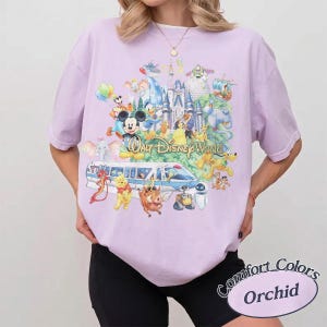 May include: A light purple short-sleeve t-shirt featuring a colorful graphic print of various Disney characters, the Walt Disney World castle, and a monorail train. The text Walt Disney World is printed in the center of the graphic design.