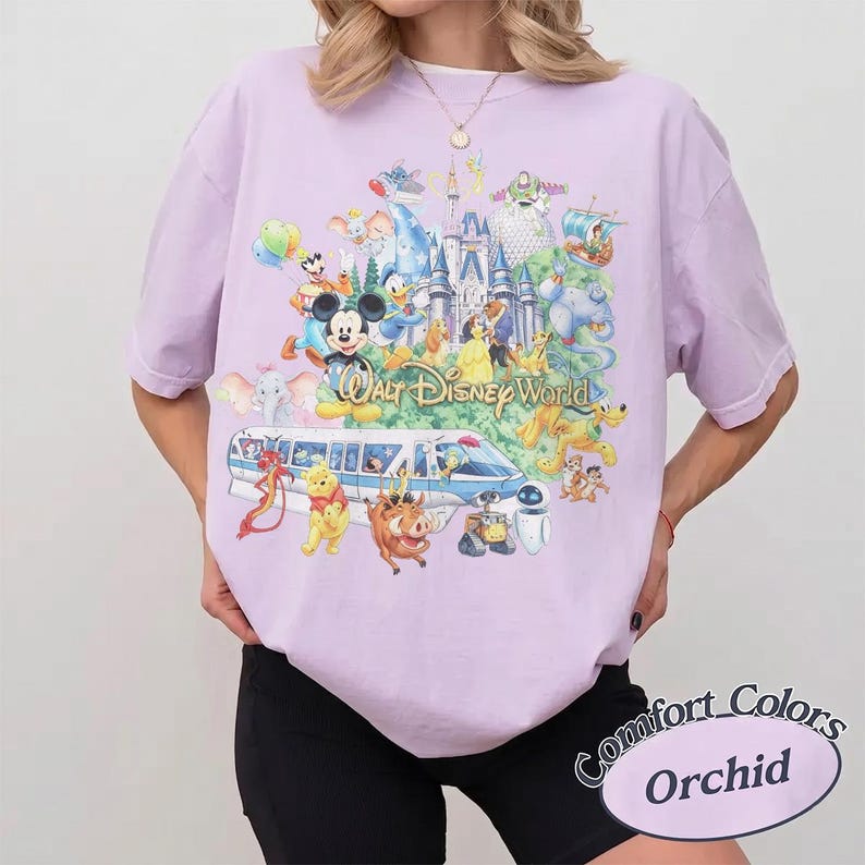 May include: A light purple short-sleeve t-shirt featuring a colorful graphic print of various Disney characters, the Walt Disney World castle, and a monorail train. The text Walt Disney World is printed in the center of the graphic design.