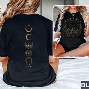 May include: Black t-shirt with gold graphic designs. The front features text that reads "VELARIS CITY OF STARLIGHT" with a mountain and star design. The back has a vertical design of crescent moons and other celestial symbols.