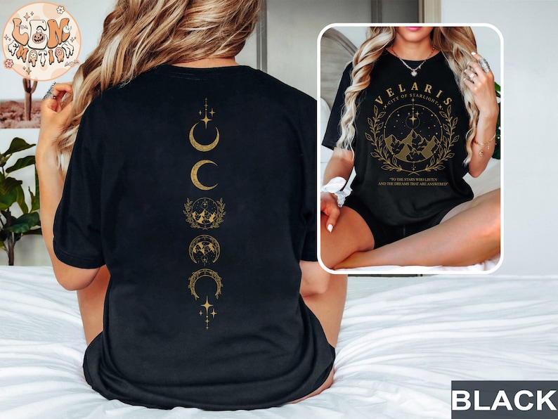 May include: Black t-shirt with gold graphic designs. The front features text that reads "VELARIS CITY OF STARLIGHT" with a mountain and star design. The back has a vertical design of crescent moons and other celestial symbols.