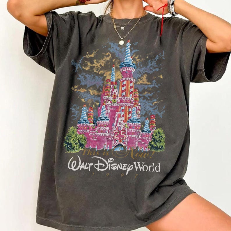 May include: Dark gray t-shirt featuring a colorful illustration of a castle with the text "This is Now! Walt Disney World 1997". The castle is pink with blue and red accents, and the number "25" is prominently displayed.