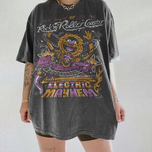 Puede incluir: Camiseta gris oscuro con un dise&ntilde;o gr&aacute;fico que presenta un personaje de los Muppets y el texto "Rock'n Roller Coaster" y "Electric Mayhem". El dise&ntilde;o incluye estrellas y elementos musicales en tonos amarillos, morados y naranjas.