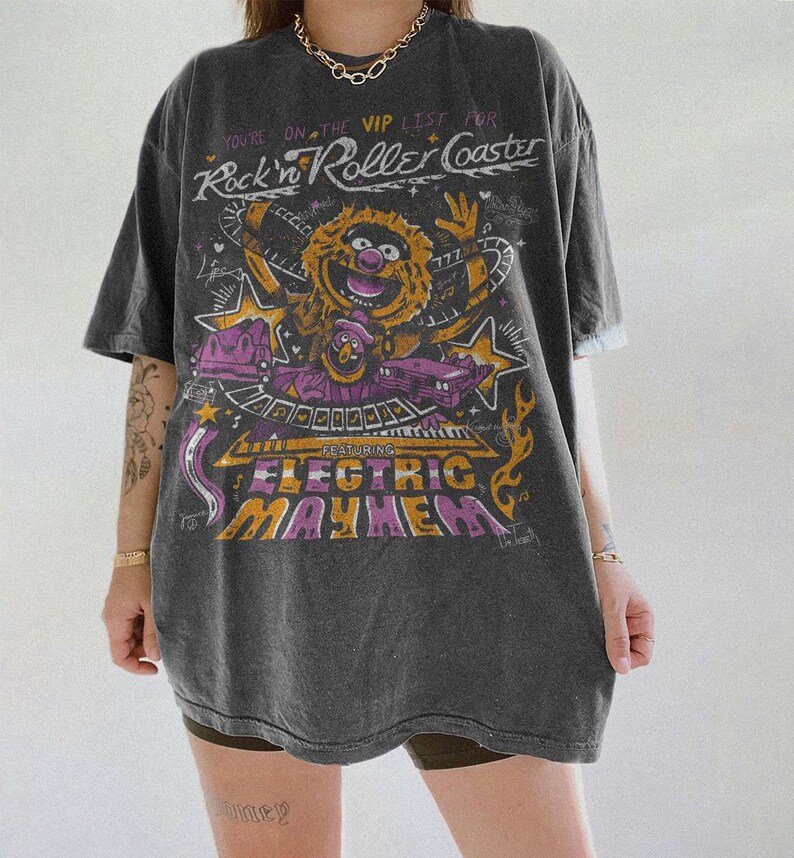 Puede incluir: Camiseta gris oscuro con un dise&ntilde;o gr&aacute;fico que presenta un personaje de los Muppets y el texto "Rock'n Roller Coaster" y "Electric Mayhem". El dise&ntilde;o incluye estrellas y elementos musicales en tonos amarillos, morados y naranjas.