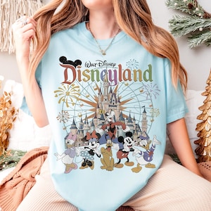May include: Light blue t-shirt featuring the Disneyland logo and various Disney characters, including Mickey Mouse, Minnie Mouse, Donald Duck, and Goofy. The design includes a castle and fireworks.