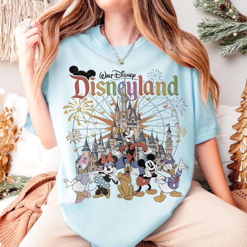 May include: Light blue t-shirt featuring the Disneyland logo and various Disney characters, including Mickey Mouse, Minnie Mouse, Donald Duck, and Goofy. The design includes a castle and fireworks.