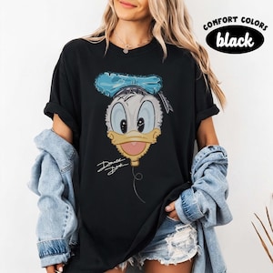 Pode incluir: Uma t-shirt preta de manga curta com um gr&aacute;fico da cabe&ccedil;a do Pato Donald desenhada como um bal&atilde;o com um la&ccedil;o azul, um bico amarelo e um fio que desce. O texto "Donald Duck" est&aacute; escrito em letra cursiva abaixo do gr&aacute;fico.
