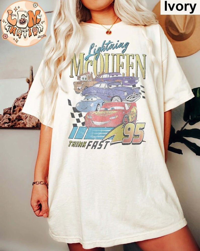 May include: A white t-shirt with a vintage-style graphic of Lightning McQueen from the Cars movie. The graphic features the text "Lightning McQueen" and "Think Fast" along with a checkered flag and the number 95. The shirt is a light ivory color.
