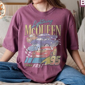 May include: A purple t-shirt with a graphic print of Lightning McQueen and other Cars characters. The text "Lightning McQueen", "Think Fast", and "95" are printed on the shirt.
