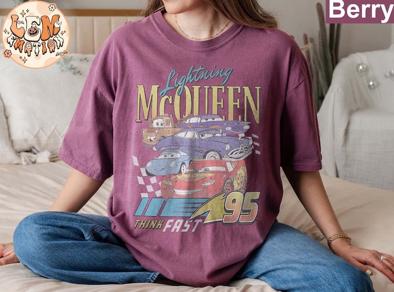 May include: A purple t-shirt with a graphic print of Lightning McQueen and other Cars characters. The text "Lightning McQueen", "Think Fast", and "95" are printed on the shirt.