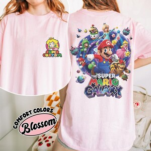 Pu&ograve; includere: T-shirt rosa chiaro con grafica Super Mario Galaxy sul retro e logo Principessa Peach sul davanti. La grafica sul retro include Mario, Luigi e altri personaggi. La maglietta ha il testo "Super Custom" e "Comfort Colors Blossom".