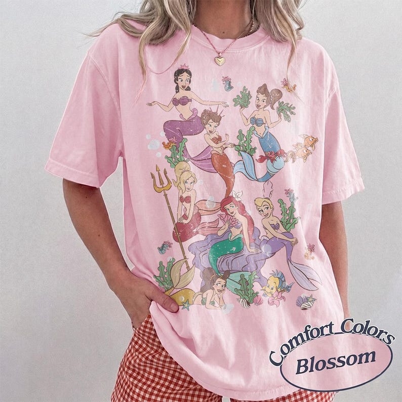 Puede incluir: Una camiseta de manga corta de color rosa claro con un estampado gr&aacute;fico de varias sirenas de dibujos animados con diversos colores de pelo y dise&ntilde;os de cola, rodeadas de algas y burbujas. El texto Comfort Colors Blossom aparece abajo.