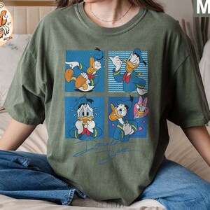 May include: A green t-shirt with a graphic print of Donald Duck in different poses. The shirt has a crew neck and short sleeves. The text "Donald Duck" is printed on the bottom of the shirt.