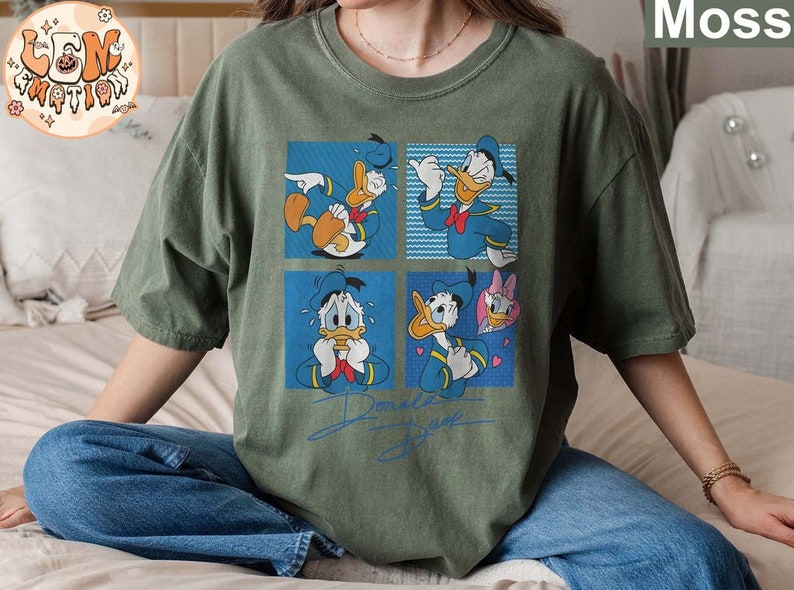 May include: A green t-shirt with a graphic print of Donald Duck in different poses. The shirt has a crew neck and short sleeves. The text "Donald Duck" is printed on the bottom of the shirt.