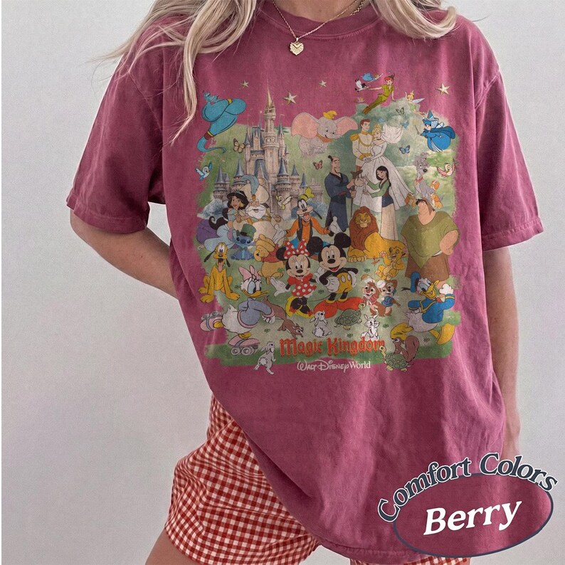 May include: A berry-colored short-sleeve t-shirt featuring a large, colorful graphic print of various Disney characters and a castle. The text "Magic Kingdom" and "Walt Disney World" is printed in red and white below the character illustration.