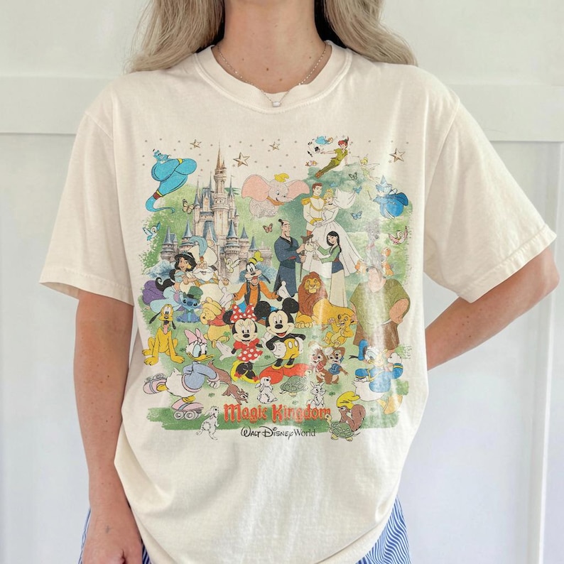 May include: A cream-colored short-sleeve t-shirt featuring a colorful graphic print of various Disney characters, including Mickey Mouse, Minnie Mouse, and Cinderella, in front of a castle. The text "Magic Kingdom" and "Walt Disney World" is printed below.
