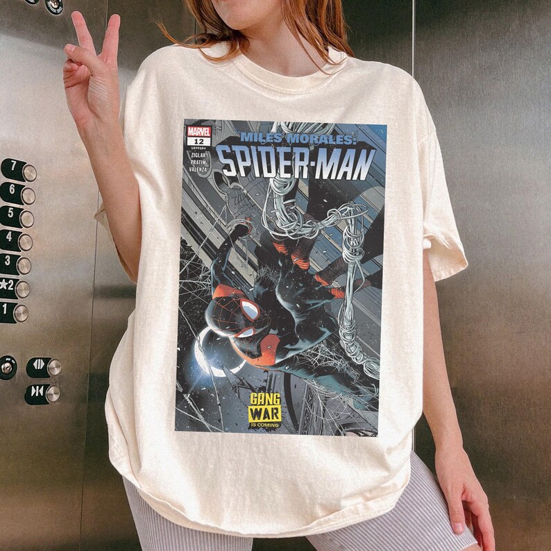 Op de afbeelding: Cr&egrave;mekleurig T-shirt met een stripboekcover van Miles Morales: Spider-Man. De cover heeft een zwart-rode Spider-Man illustratie met de tekst "GANG WAR IS COMING". Het Marvel-logo en nummer 12 zijn ook zichtbaar.