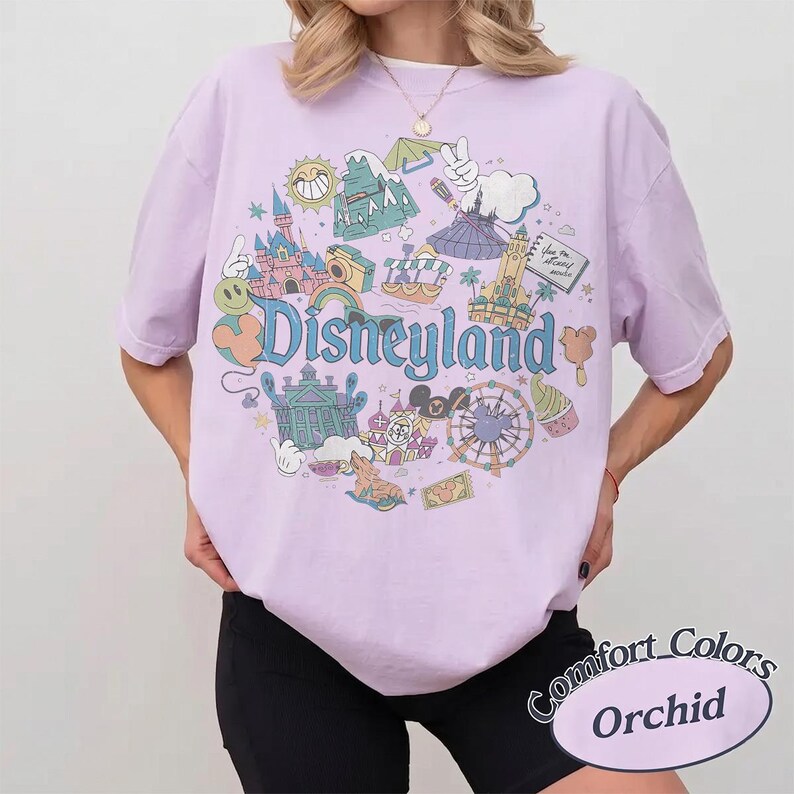May include: A light purple t-shirt with a Disneyland graphic. The graphic features various park icons, including a castle, Matterhorn, and a Ferris wheel, with the word "Disneyland" in a whimsical font. The shirt is labeled "Comfort Colors Orchid".