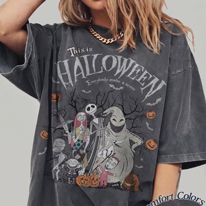 Pu&ograve; includere: T-shirt grigio scuro con grafica di "Nightmare Before Christmas". Il design include personaggi come Jack Skellington, Sally e Oogie Boogie, con la scritta "This is Halloween". La maglietta &egrave; Comfort Colors Pepper.