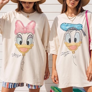Pode incluir: Duas t-shirts oversize de cor creme com estampados gr&aacute;ficos de rostos de patos de desenhos animados em forma de bal&atilde;o. A t-shirt da esquerda mostra a Daisy Duck com um la&ccedil;o cor-de-rosa e o texto Daisy Duck. A da direita mostra o Donald Duck com um chap&eacute;u azul e o texto Donald Duck.