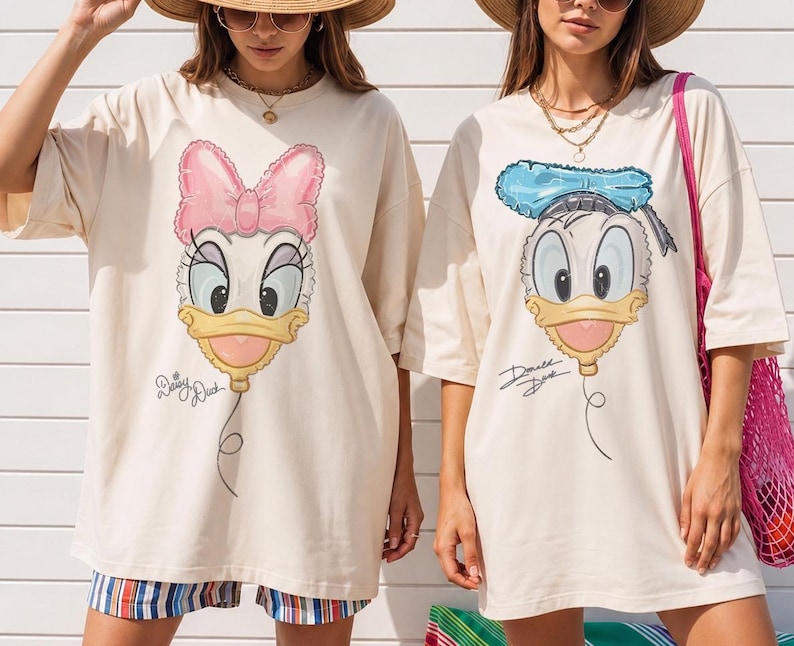 Pode incluir: Duas t-shirts oversize de cor creme com estampados gr&aacute;ficos de rostos de patos de desenhos animados em forma de bal&atilde;o. A t-shirt da esquerda mostra a Daisy Duck com um la&ccedil;o cor-de-rosa e o texto Daisy Duck. A da direita mostra o Donald Duck com um chap&eacute;u azul e o texto Donald Duck.