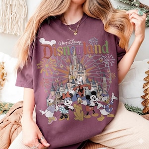 May include: A maroon t-shirt featuring a colorful graphic of Disneyland characters and the Disneyland logo. The graphic includes Mickey Mouse, Minnie Mouse, Donald Duck, and Goofy in front of a castle and fireworks.