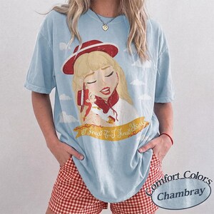 Pode incluir: Uma t-shirt de manga curta azul-clara com uma estampa gr&aacute;fica de uma personagem loira usando um chap&eacute;u vermelho e segurando uma boneca pequena. Abaixo da ilustra&ccedil;&atilde;o, h&aacute; uma faixa amarela com o texto I Knew It, I Knew It em cursivo.