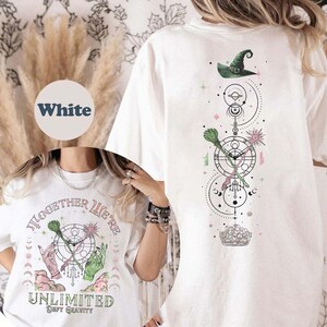 May include: White t-shirt with a graphic design of a witch's hat, broomsticks, and a crown. The text "Together We're Unlimited" and "Defy Gravity" is printed on the shirt.