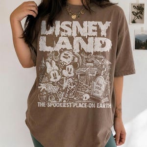 May include: Brown t-shirt with a distressed "Disney Land" graphic. The graphic features Mickey and Minnie Mouse in skeleton costumes, a graveyard scene, and the text "The Spookiest Place on Earth."