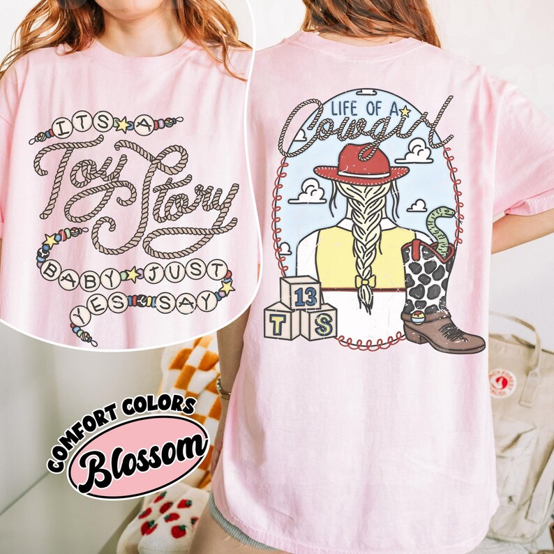 May include: A light pink t-shirt featuring a graphic on the back of a cowgirl with a braided ponytail, a red hat, and a cow-print boot with a snake. The text reads "LIFE OF A COWGIRL" and "IT'S A TOY STORY BABY JUST SAY YES". Includes alphabet blocks.
