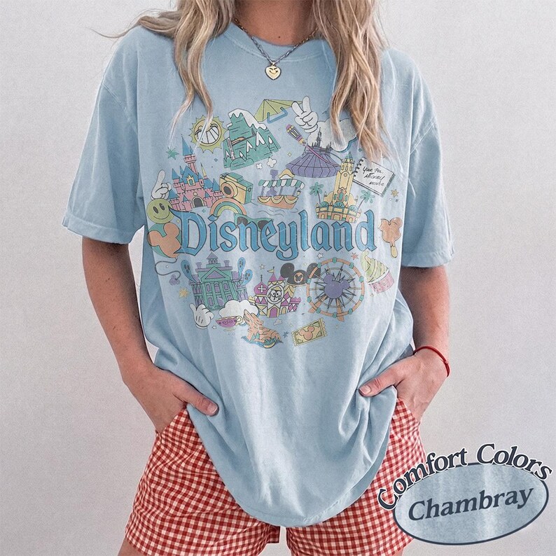May include: Light blue t-shirt with a Disneyland graphic. The graphic features various park icons and the word "Disneyland" in a whimsical font. The shirt is paired with red and white checkered shorts.