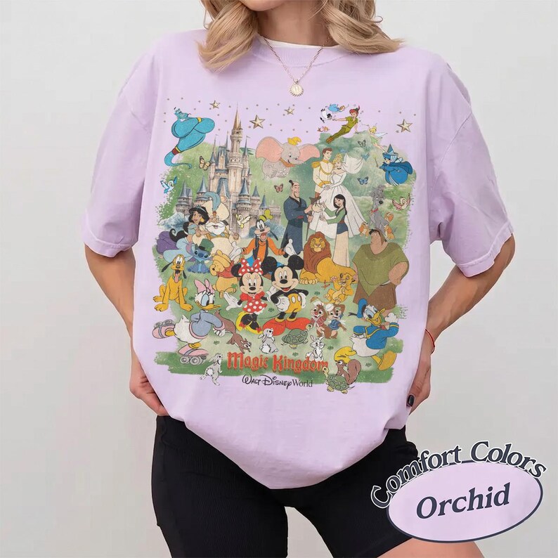 May include: A light purple t-shirt featuring a large graphic print of various Disney characters, including Mickey Mouse, Minnie Mouse, and Cinderella, in front of a castle. The text "Magic Kingdom" and "Walt Disney World" is printed below the characters.