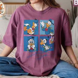 May include: A purple t-shirt with a graphic print of Donald Duck in four different poses. The shirt has a crew neck and short sleeves.
