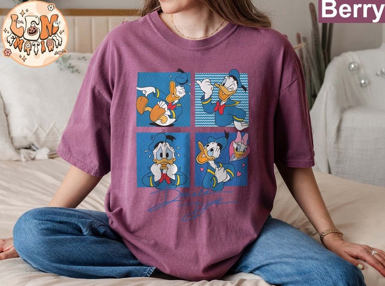 May include: A purple t-shirt with a graphic print of Donald Duck in four different poses. The shirt has a crew neck and short sleeves.