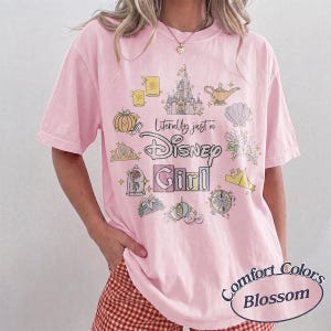 Op de afbeelding: Roze T-shirt met de tekst "Literally just a Disney Girl" en Disney-thema illustraties. Het shirt heeft een ronde hals en korte mouwen. Het shirt is een Comfort Colors Blossom.