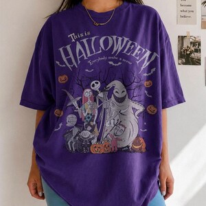 Pu&ograve; includere: Una maglietta viola con una grafica del film "Nightmare Before Christmas". Il design include Jack Skeletron, Sally e altri personaggi, con la scritta "This is Halloween" e "Everybody make a scene."