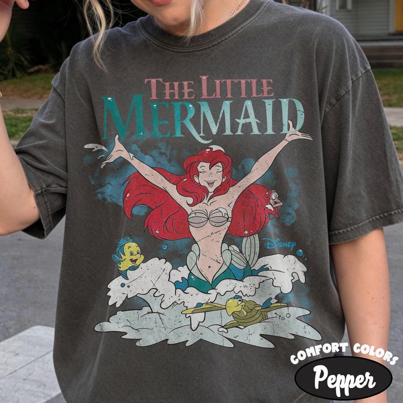 May include: A charcoal gray t-shirt featuring a graphic of Ariel from The Little Mermaid with red hair, arms outstretched, surrounded by Flounder and Sebastian. The text reads The Little Mermaid in teal and red, with a small Disney logo included.