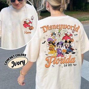 May include: Ivory-colored long-sleeve t-shirt with a Halloween-themed Disney design. The front features a cartoon Donald Duck with a devil's costume. The back displays Disney characters in Halloween costumes with the text "Disneyworld Florida Est 1971".