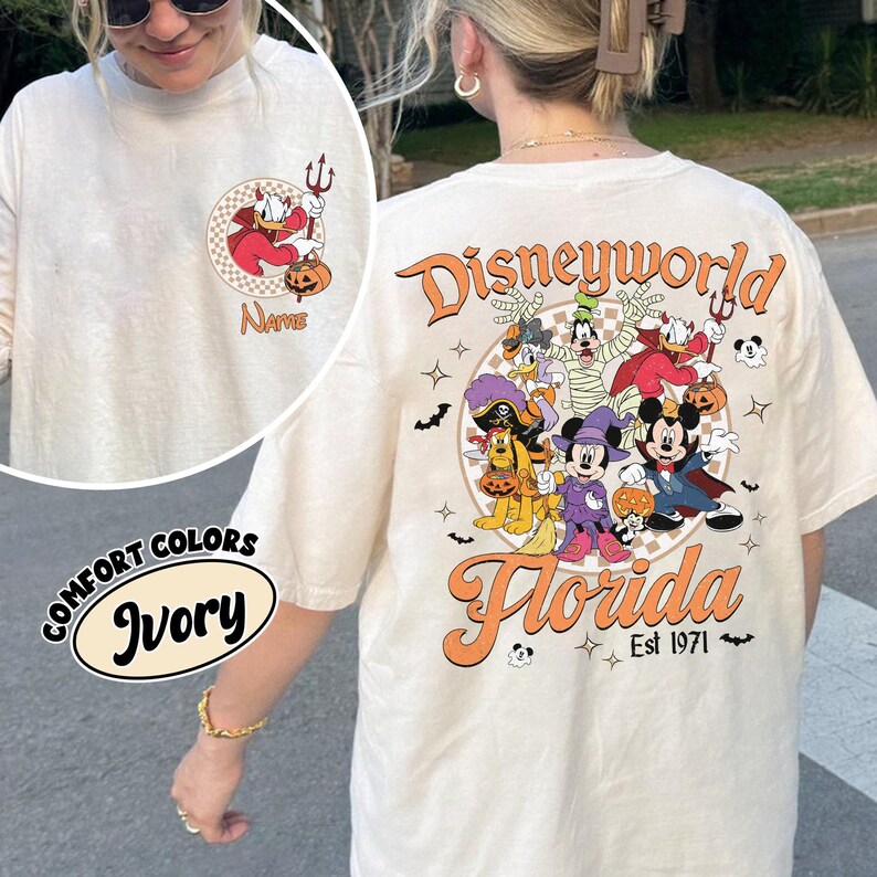 May include: Ivory-colored long-sleeve t-shirt with a Halloween-themed Disney design. The front features a cartoon Donald Duck with a devil's costume. The back displays Disney characters in Halloween costumes with the text "Disneyworld Florida Est 1971".
